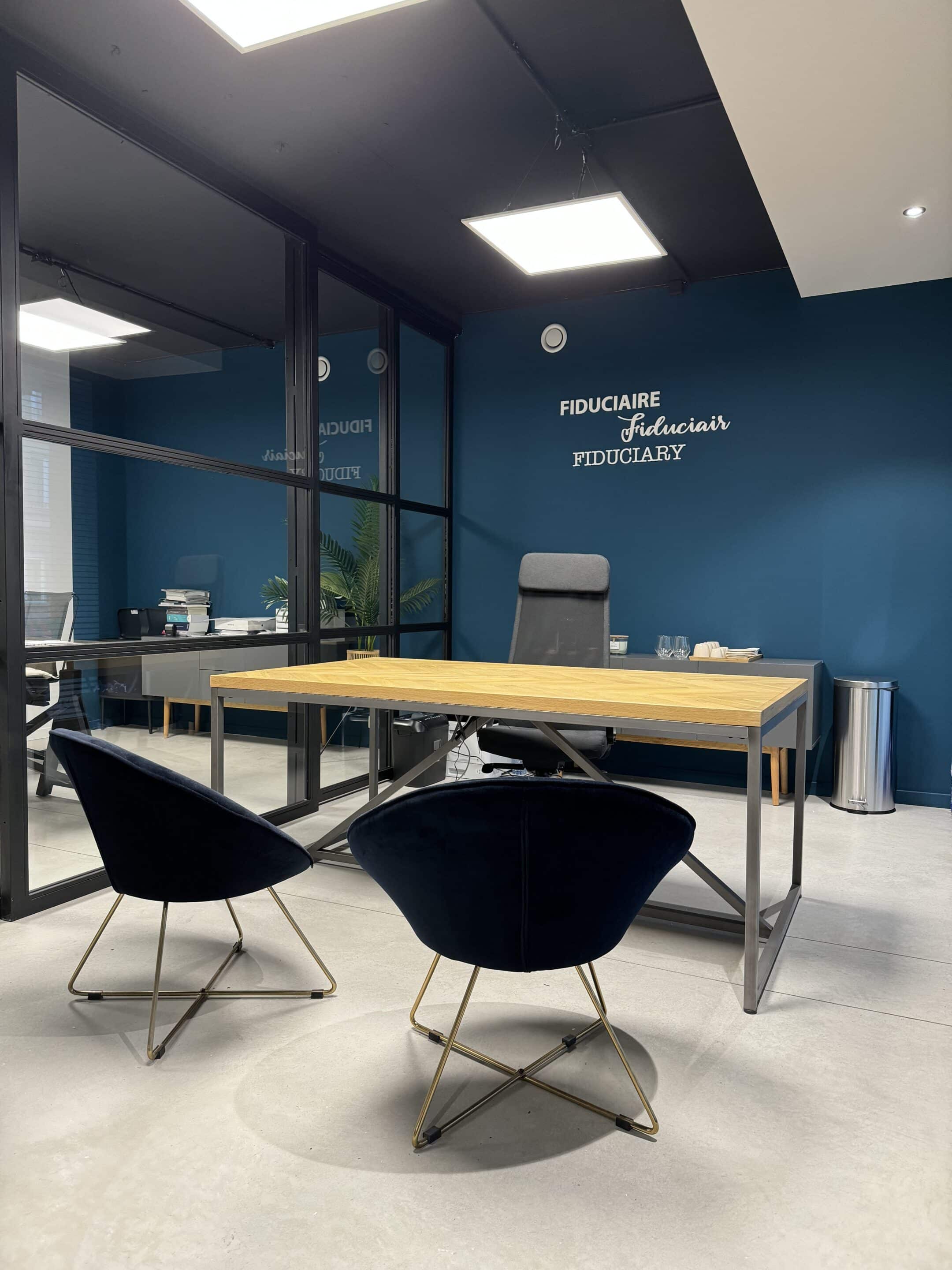 Photo d'un bureau moderne avec une table en bois, deux chaises noires et un mur bleu portant les inscriptions 'FIDUCIAIRE' AAM-Solutions Domiciliation d'entreprises Bruxelles