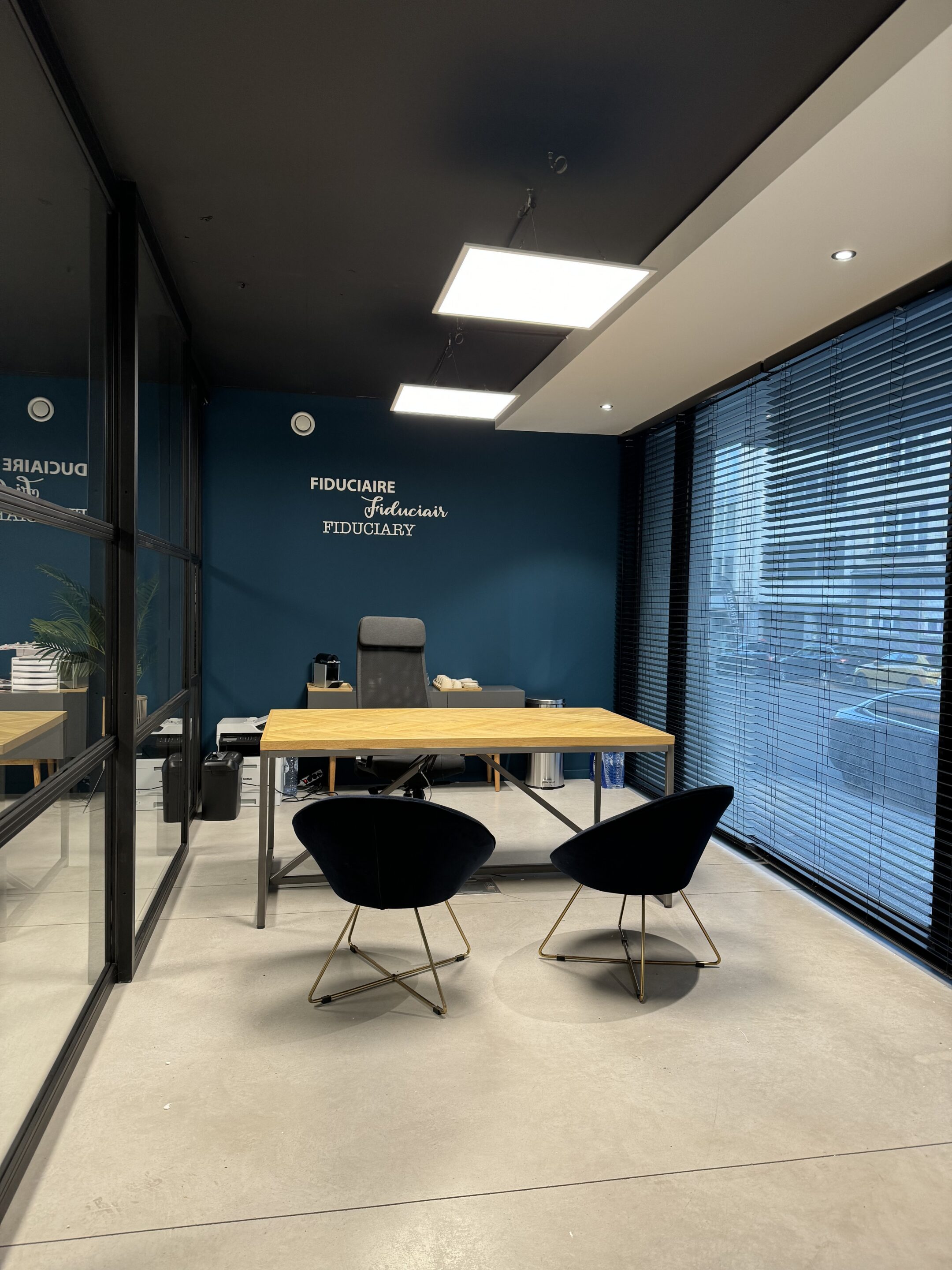 Photo d'un bureau moderne avec une table en bois, deux chaises noires et un mur bleu portant les inscriptions 'FIDUCIAIRE' AAM-Solutions Domiciliation d'entreprises et siège social Bruxelles La Cambre